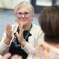 Woman sitting at table, clapping and looking at camera while listening to speaker